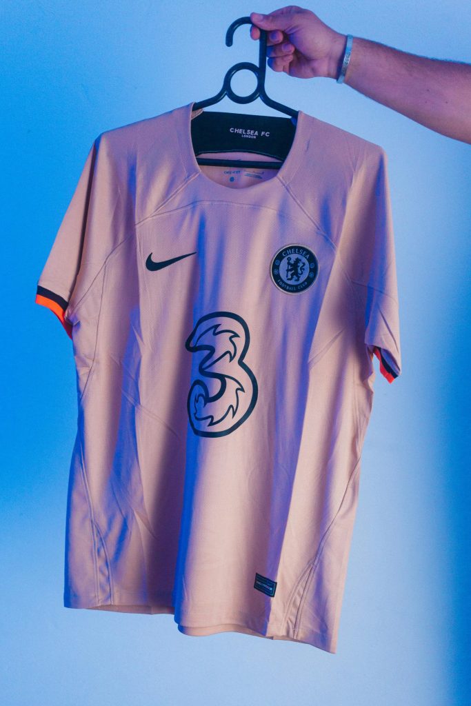 Chelsea FC soccer jersey displayed on a hanger against a blue background, featuring Nike logo and sponsor.